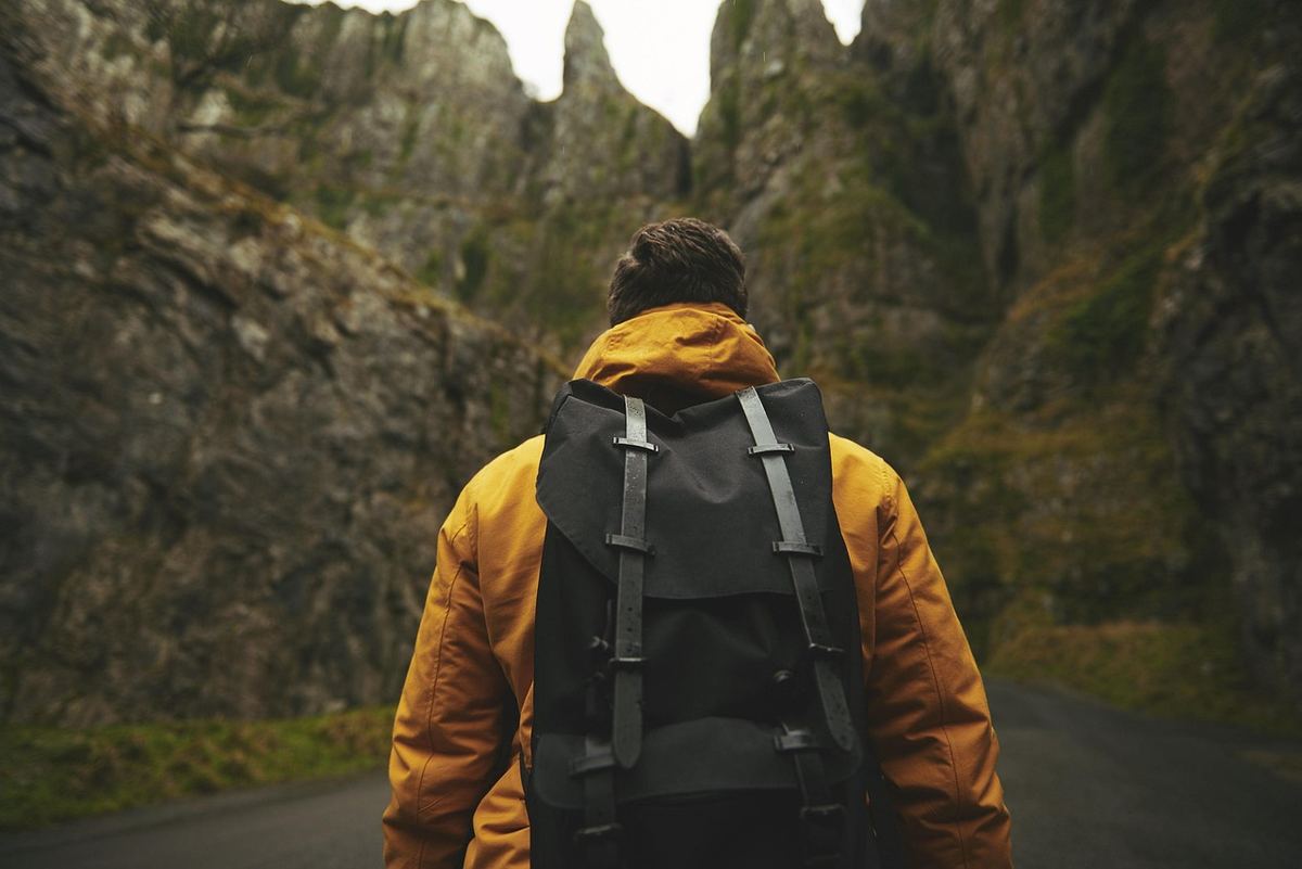 Homme en manteau jaune marche sur une route avec un sac noir, entouré de montagnes rocheuses et de végétation
